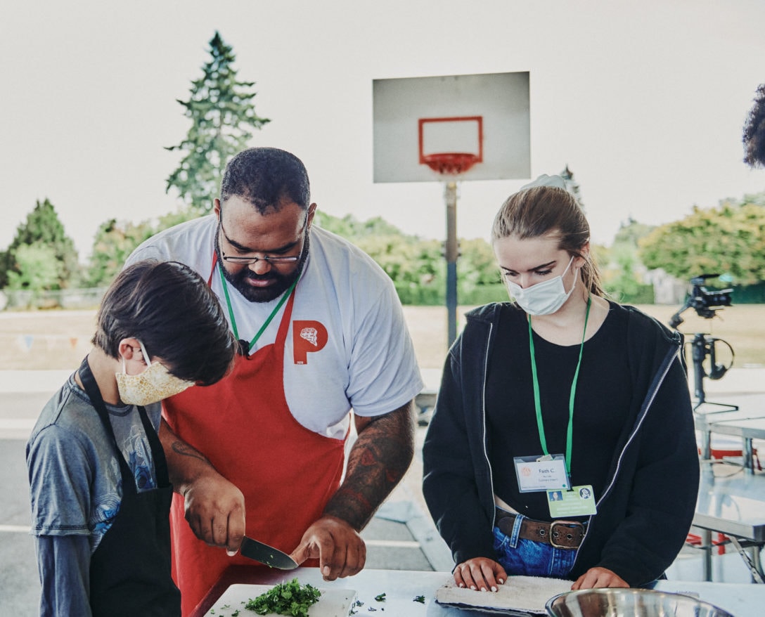 Adult shows one boy and one girl how to chop vegetables on table outside in front of a basketball hoop